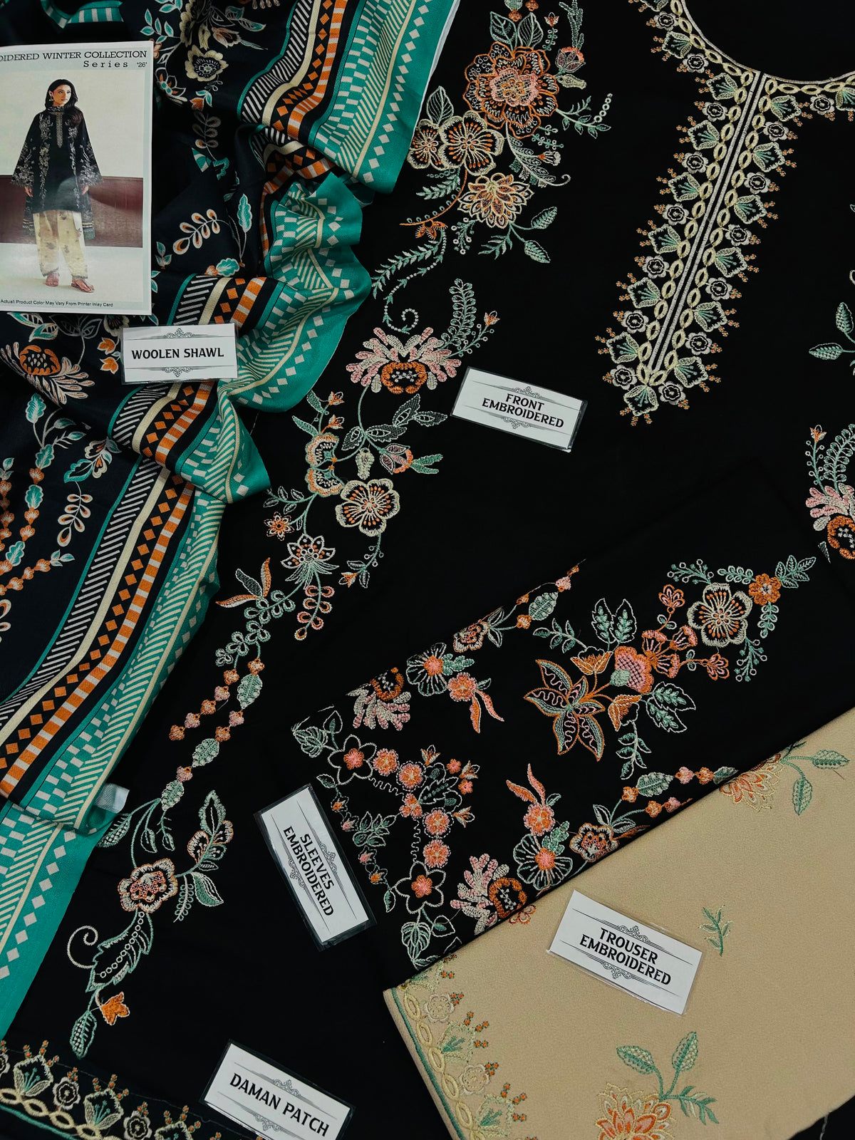 A styled flat-lay of embroidered black fabric garments with floral and vine patterns in teal, orange, and beige, alongside a beige fabric sample card and printed fabric labels.