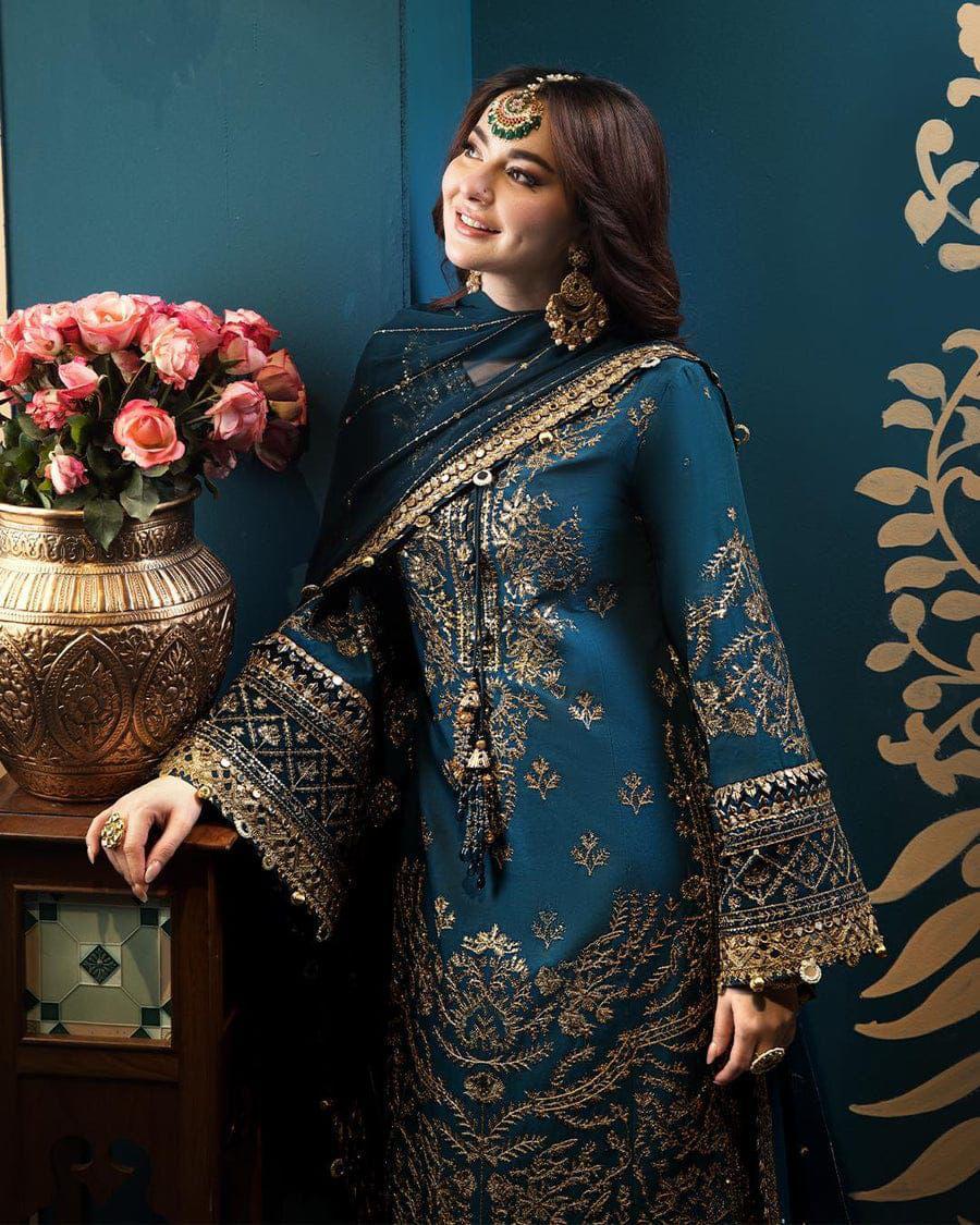 Woman in an embroidered blue and gold traditional outfit standing next to a decorative vase with flowers against a blue wall.