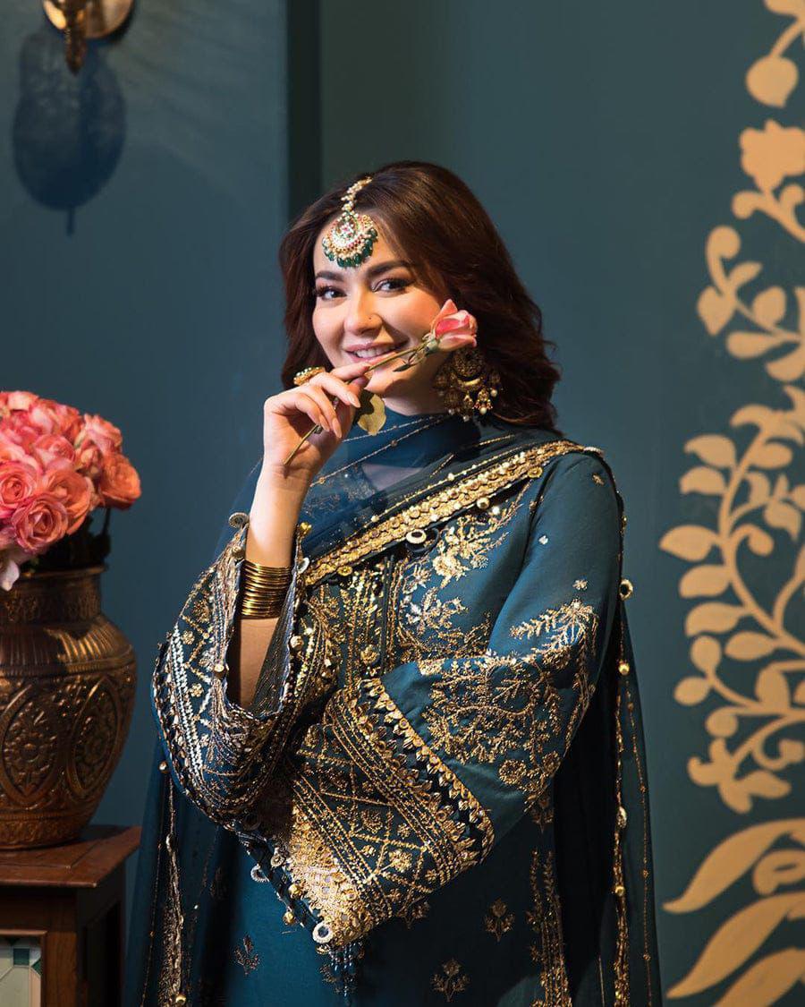 Woman in traditional embroidered outfit with floral decorations against a decorative wall.