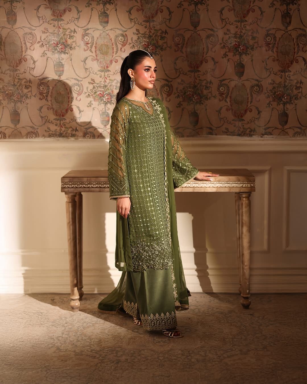 Woman in a green embroidered outfit standing in an elegant room with decorative wallpaper.