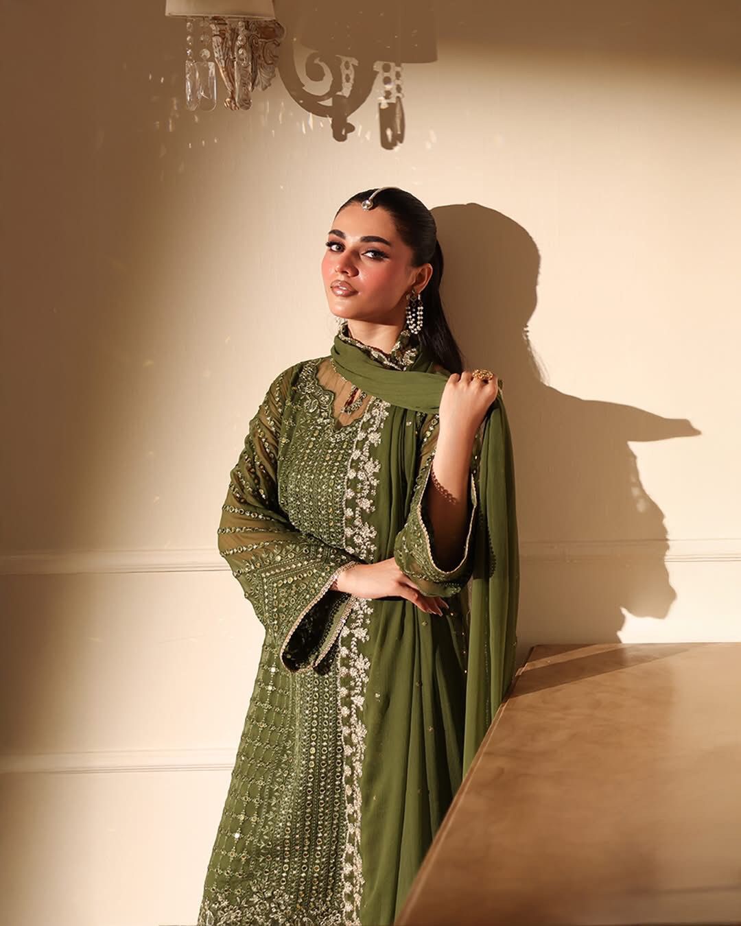 Woman in a green traditional outfit with intricate patterns, standing against a neutral wall.