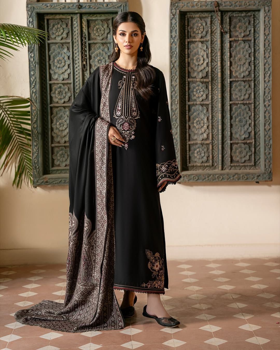 Woman in a black traditional outfit with intricate patterns standing in a decorated room.