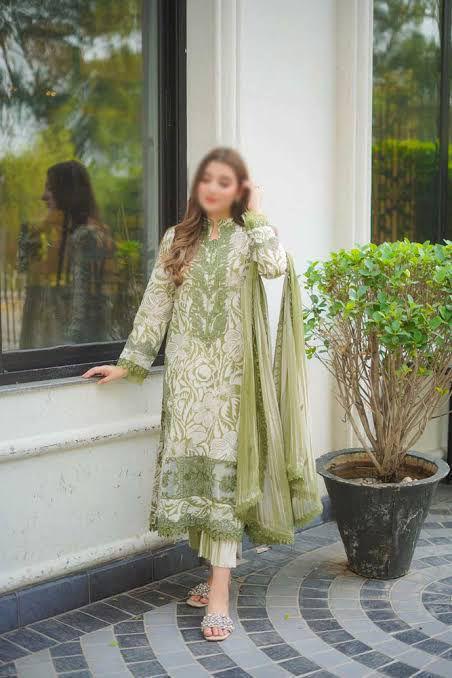 Woman in a green and white traditional outfit standing by a window with a plant in the foreground.
