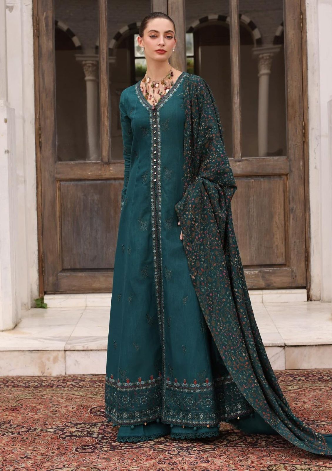 Woman in a teal traditional outfit with a long scarf standing in front of wooden doors.