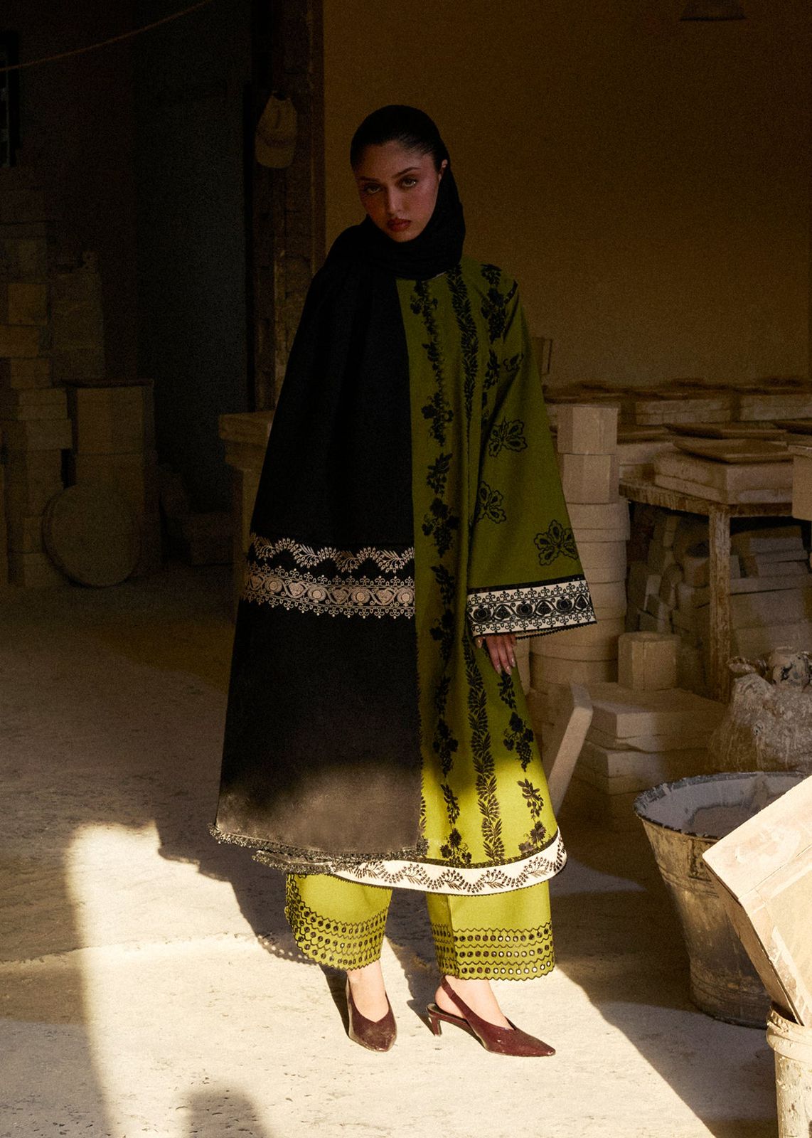 Woman in a black and green traditional outfit standing in a rustic setting with wooden barrels.