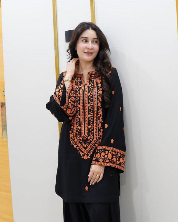 Woman wearing a black and orange embroidered traditional outfit against a white wall.