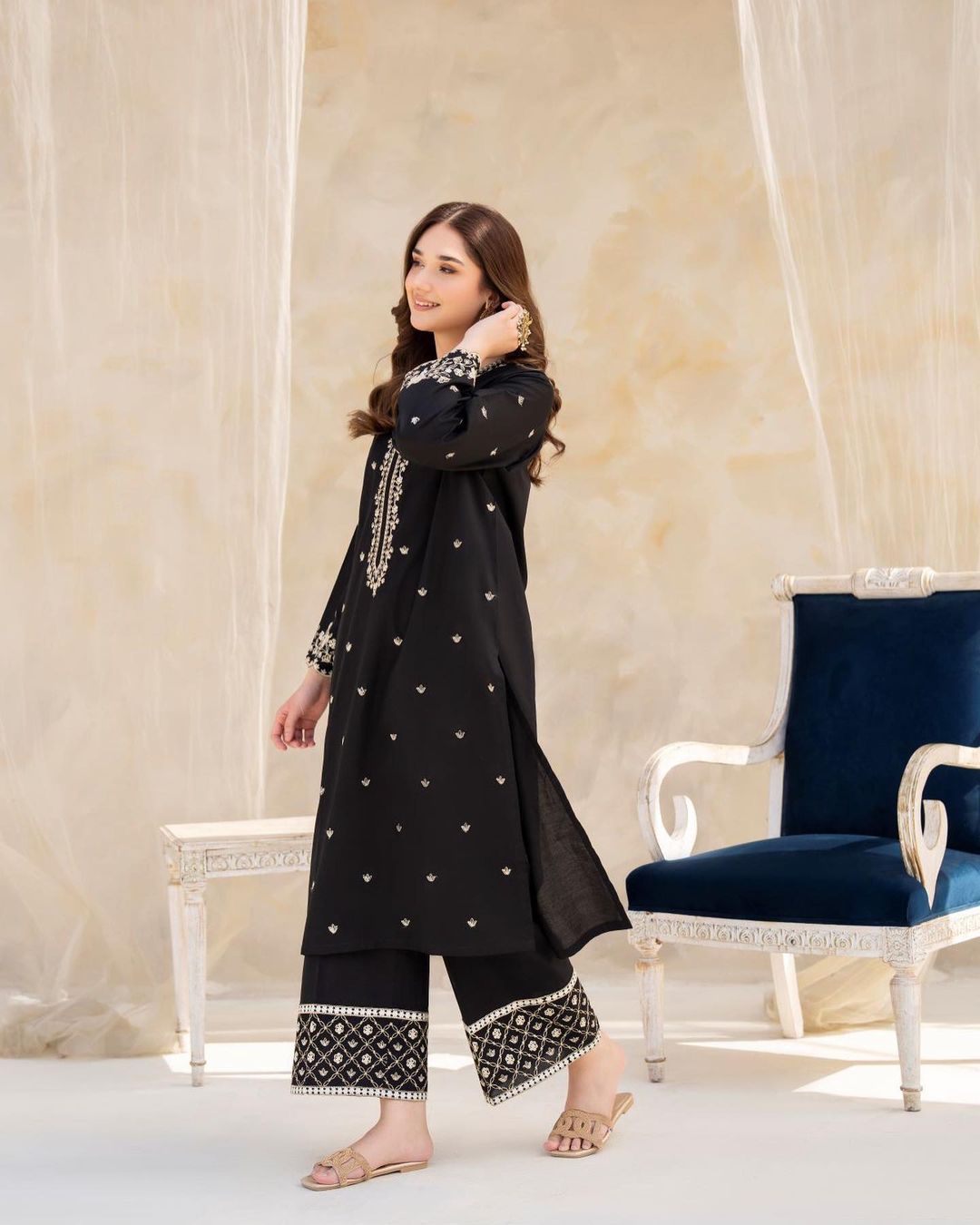Woman in a black embroidered outfit standing in a decorated room with a chair and curtains.