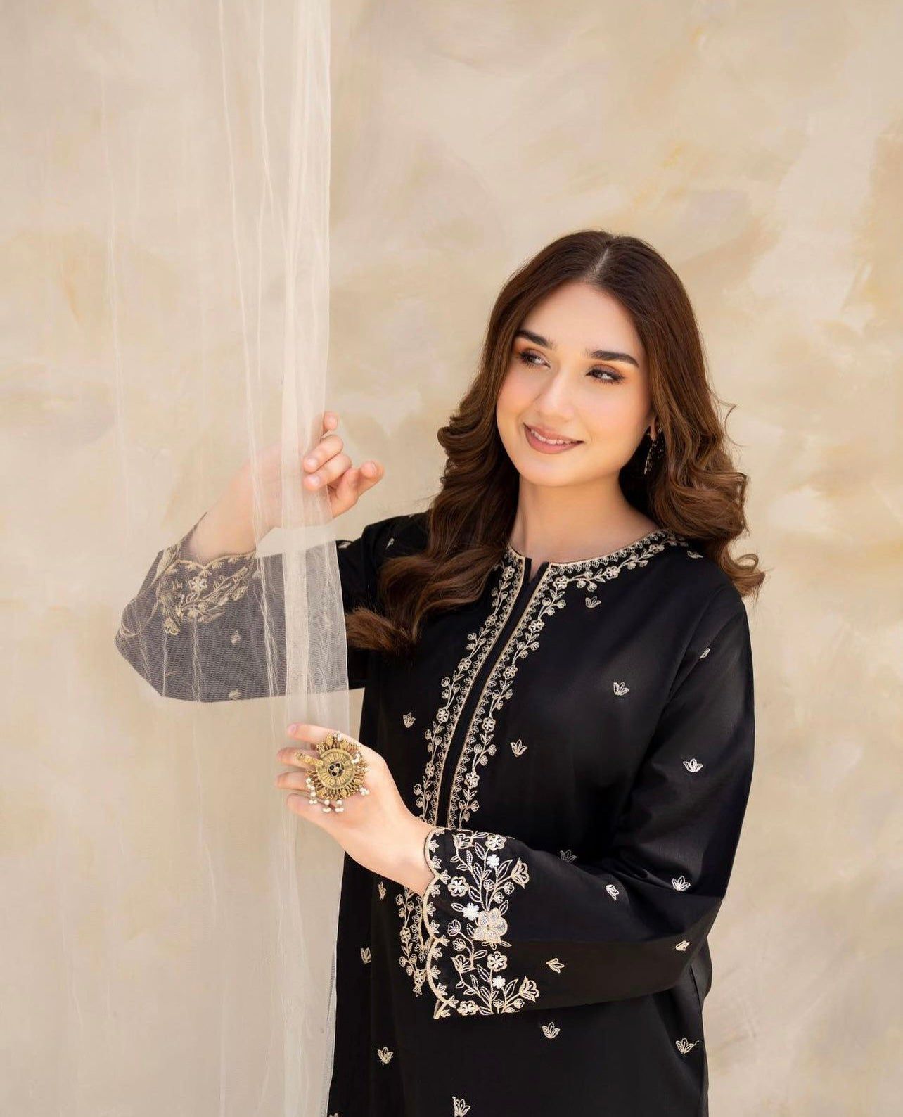 Woman in an embroidered black outfit holding a decorative item against a beige background