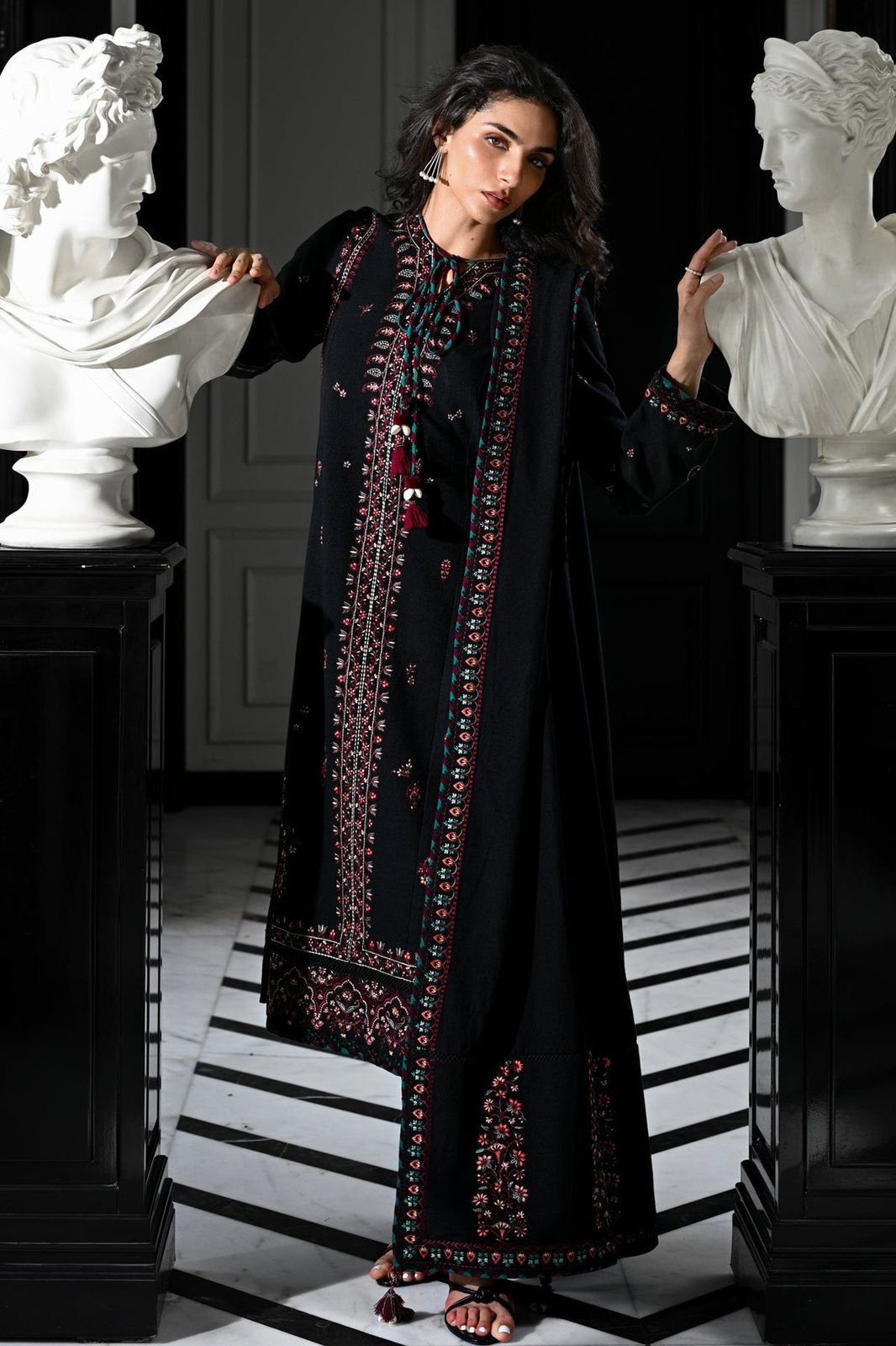 Woman in a black embroidered outfit standing between two classical busts in an elegant interior setting.