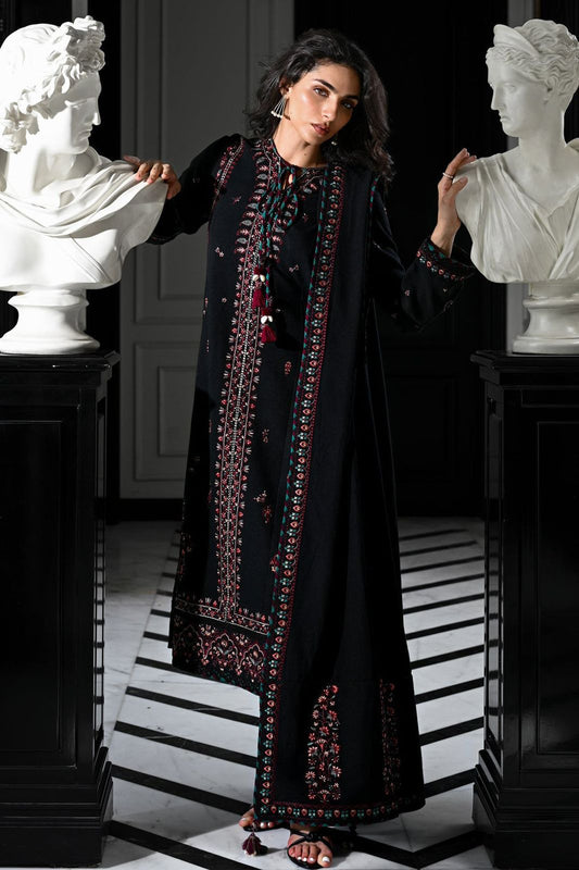 Woman in a black embroidered outfit standing between two classical busts in an elegant interior setting.