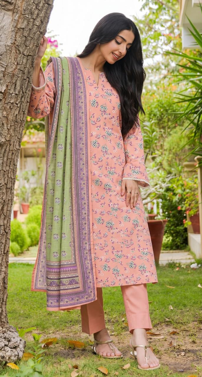A woman modeling a three-piece printed lawn collection outfit from KulAhmed Summer Collection, standing outdoors next to a tree. The outfit consists of a light pink printed kurta, matching trousers, and a green and pink patterned dupatta.