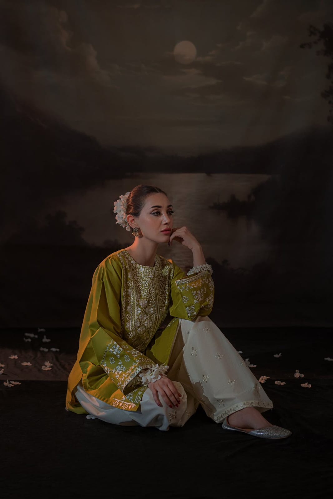 Woman in a yellow and white traditional outfit sitting by a body of water under a moonlit sky.