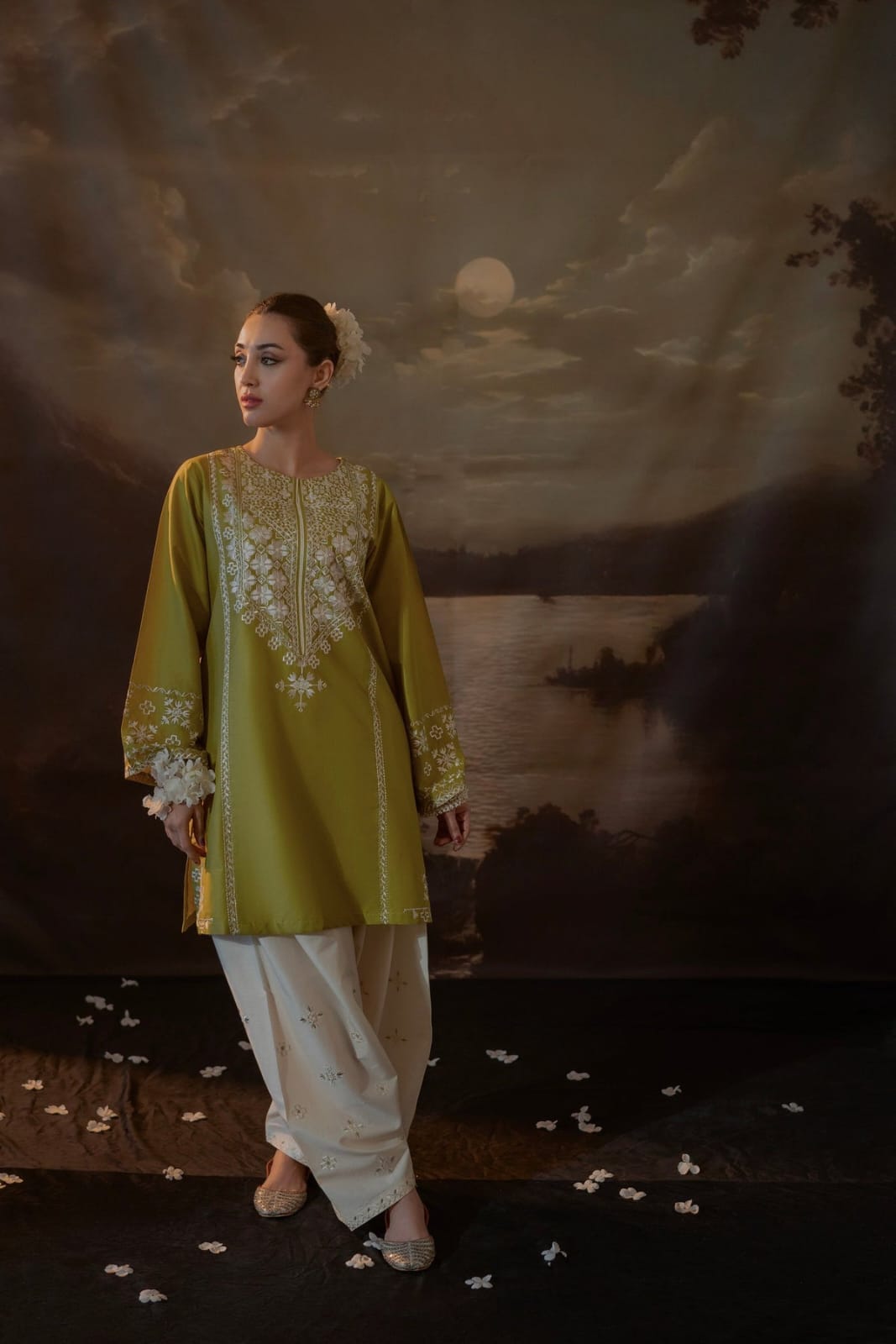 Woman in a green embroidered top and white pants standing against a painted landscape backdrop.