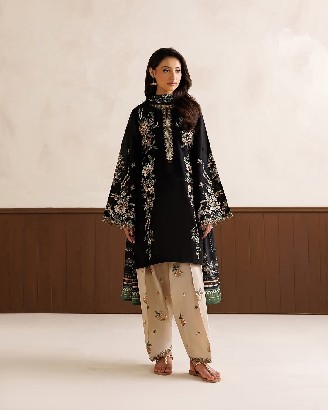 Woman wearing a black embroidered outfit with beige pants against a plain background
